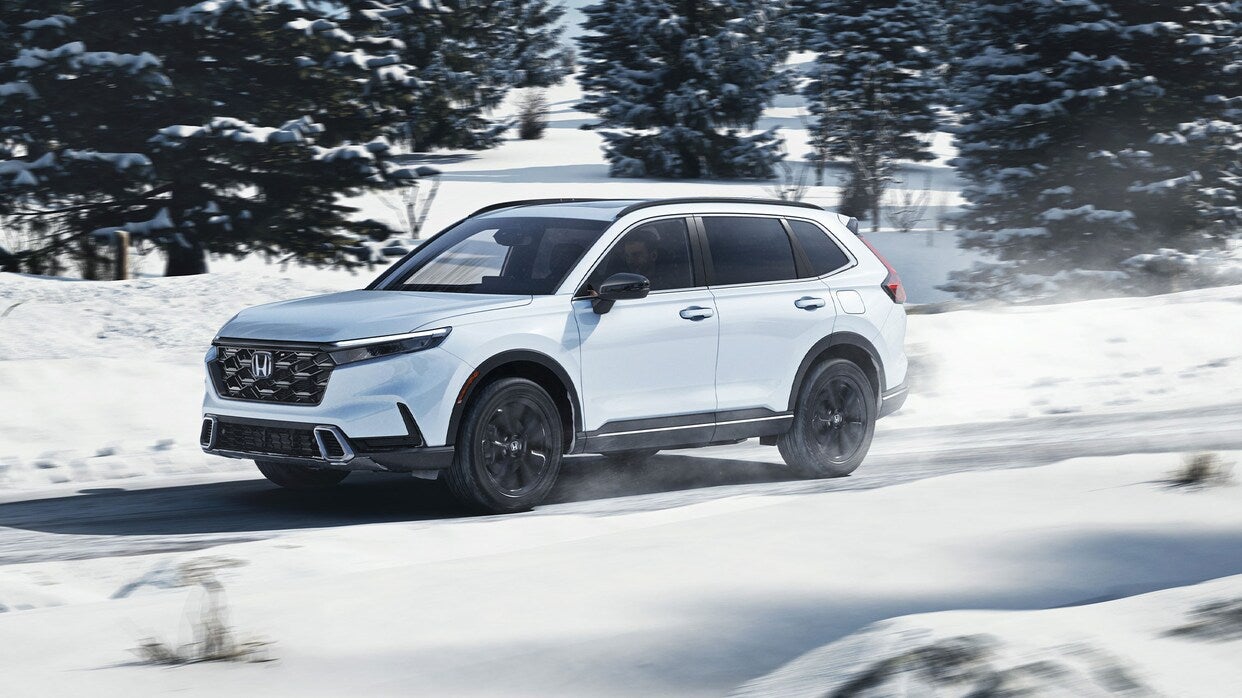 a white 2025 honda cr-v hybrid driving on a road in the snow with trees in the background