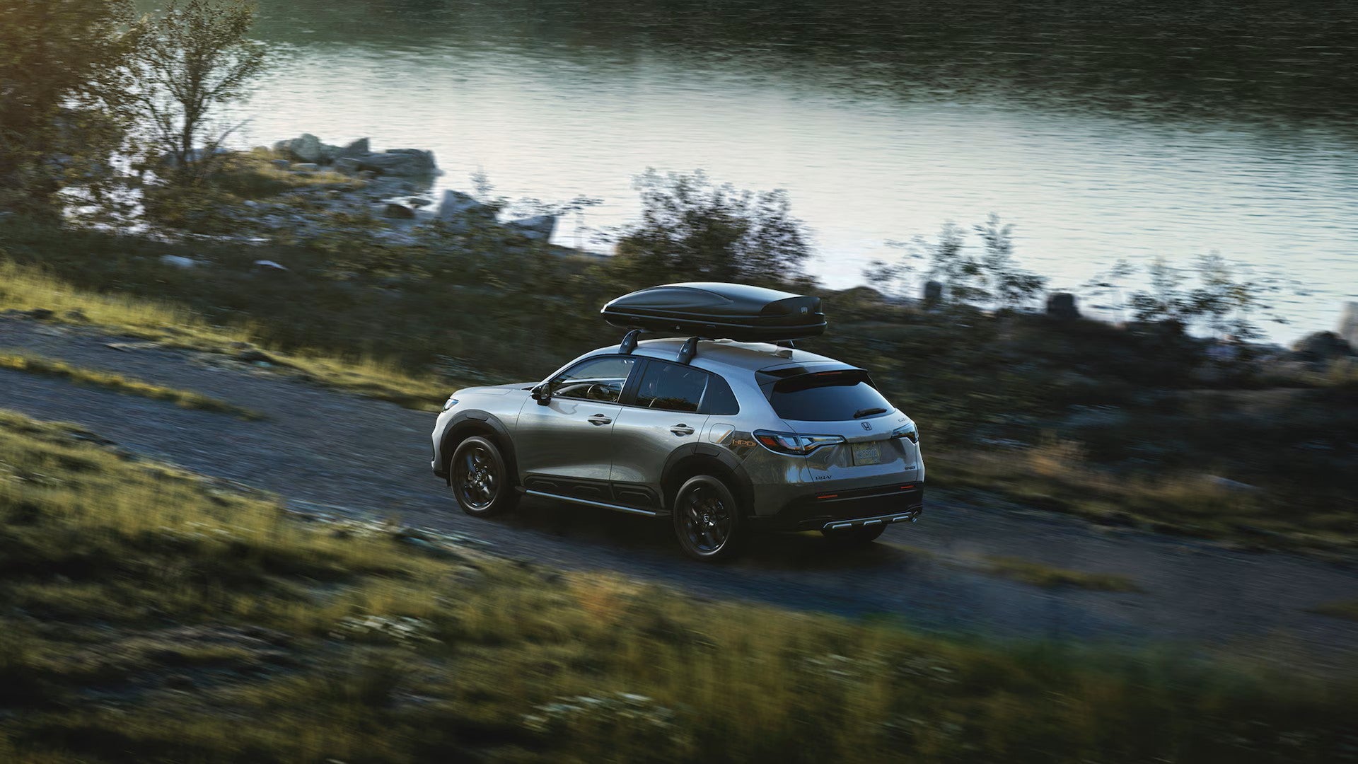 a 2025 honda hr-v driving on a gravel trail near a body of water with a luggage shell on top of the racks
