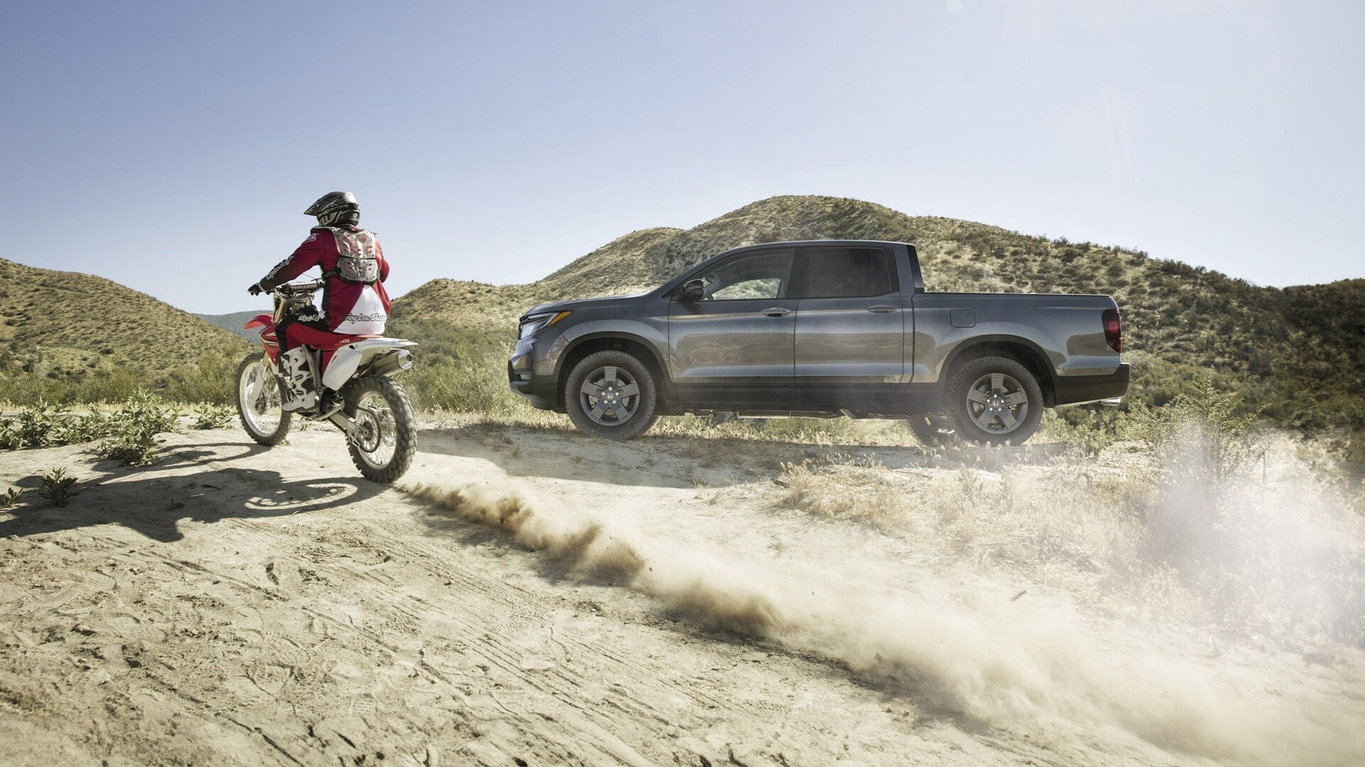2025 Honda Ridgeline Trailsport in Modern Steel Metallic with one dirt bike rider in riding gear riding across foreground.