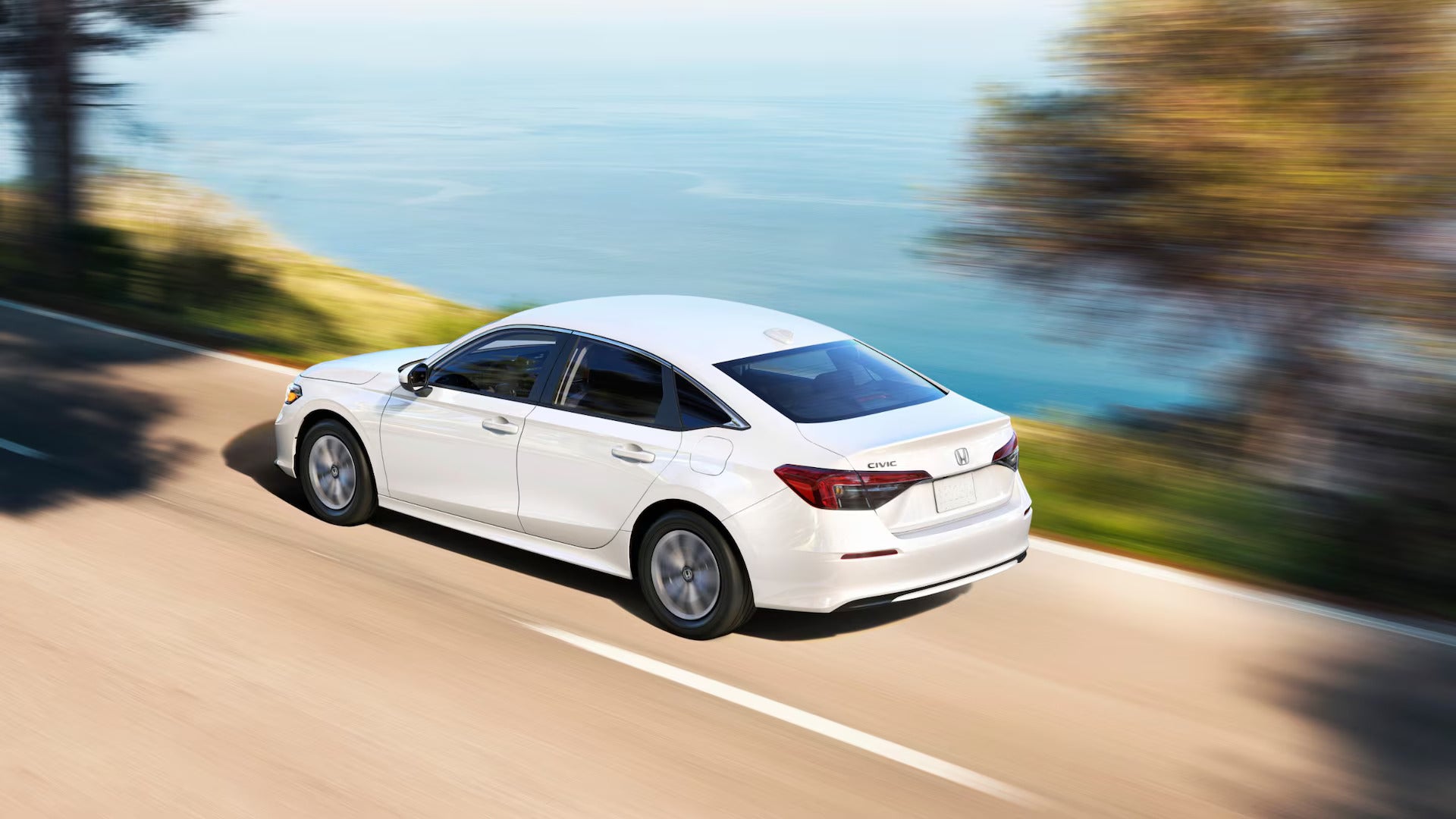 Rear side view of a white 2025 Honda Civic driving along a coastal road, showcasing its sleek design and dynamic performance.